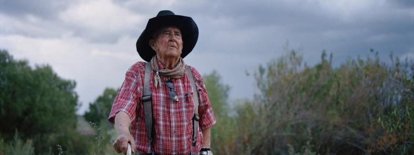 An elderly man wearing a black hat, red plaid shirt, and suspenders stands outdoors with greenery and cloudy skies in the background, looking thoughtfully into the distance.