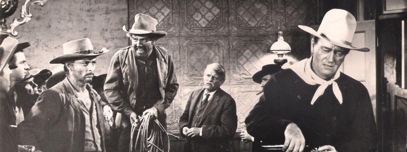 A group of men in cowboy hats and Western attire gather indoors; one man holds a coiled rope, another appears to be loading a gun, and an older man sits in the background, all in a tense black-and-white scene.