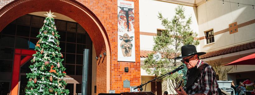 A musician in a cowboy hat plays guitar and sings outside the Autry Museum, near a large decorated Christmas tree. The scene is sunny, with festive holiday decorations and museum banners visible.