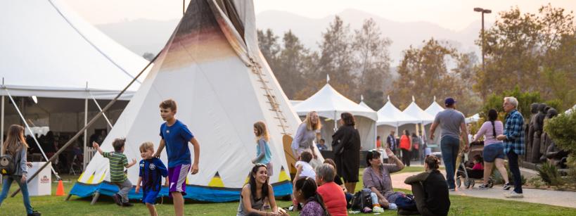 People of various ages sit and talk on the grass near a large tipi, with children playing nearby and several white tents in the background on a sunny day.
