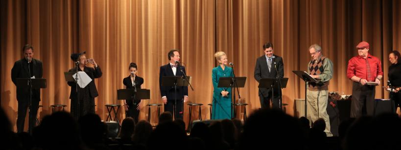 A group of people stand on stage in front of a gold curtain, each at a podium with scripts, performing a reading to an audience seated in the dark. The stage is brightly lit and the performers are dressed formally.