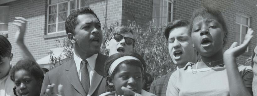 A group of young people and a man in a suit stand together outside, clapping and singing with expressive faces in front of a brick building, appearing to participate in a civil rights demonstration.