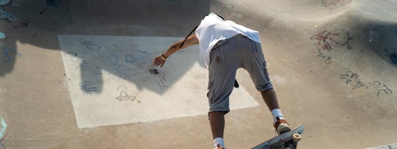 A skateboarder in a white shirt and gray shorts prepares to drop into a concrete skatepark bowl covered in graffiti and drawings, captured from behind under bright daylight.