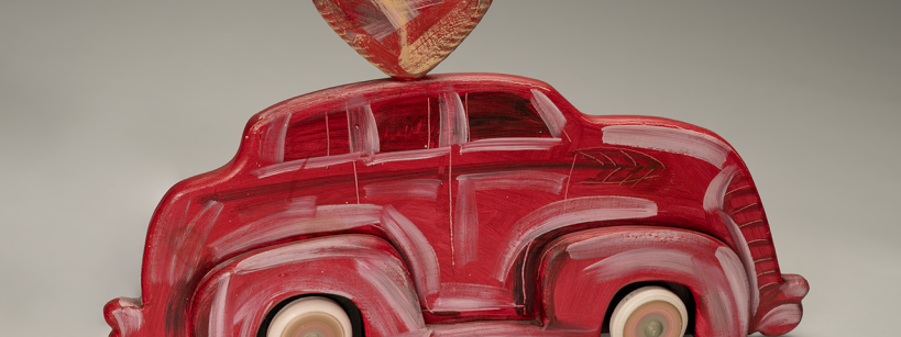 A toy car painted red with white accents, featuring a large red and gold heart shape attached to its roof, set against a plain gray background.