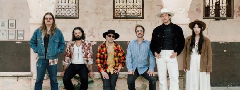 Six people stand in front of a weathered building with arched windows. They wear casual and bohemian-style clothing, including hats, sunglasses, and patterned shirts. The mood appears relaxed and vintage.