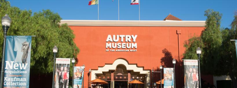 The front entrance of the Autry Museum of the American West, with banners and three flags atop the building. Tall trees and clear blue sky are visible around the museum.
