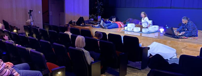People sit in theater seats watching a sound bath performance on stage, where several individuals lie down, and two facilitators are playing crystal singing bowls under blue lighting.