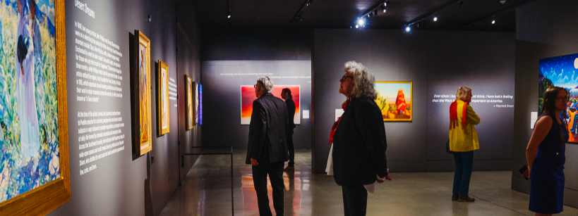 People view colorful paintings on the walls of a dimly lit art gallery. The space has polished floors, spotlights illuminating the artwork, and visitors standing and reading descriptions or admiring the art.