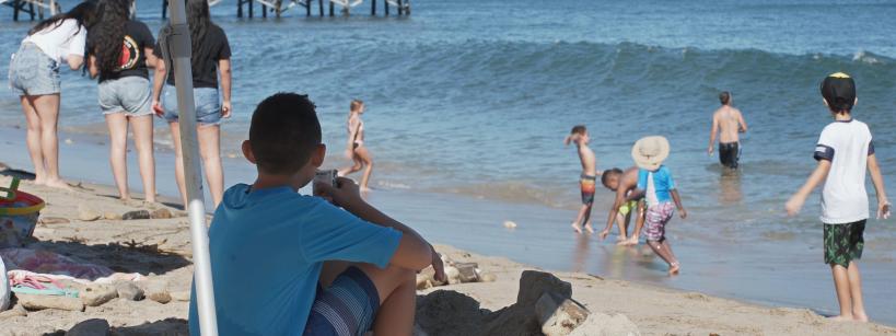Children and adults enjoy a sunny day at the beach near a pier; some play in the sand, others wade in the water, and a boy in a blue shirt sits under an umbrella, watching the scene. The ocean and hills are in the background.