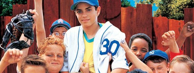 A group of smiling boys in baseball uniforms and casual clothes pose together, raising their fists in excitement. The title "The Sandlot" appears in large yellow letters above them against a bright blue sky.