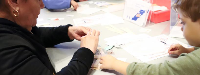 An adult and a child sit at a table working on a craft project with paper, scissors, and other materials. Other children and adults are visible in the background, also participating in the activity.