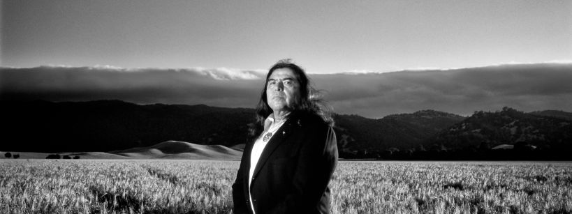 A man wearing a suit stands in a vast field of tall grass, with rolling hills and a cloudy sky in the background, captured in black and white.