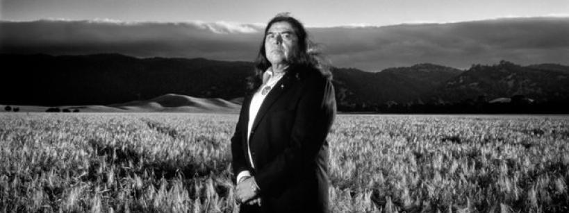 A man with long hair, dressed in a suit, stands in a field of tall grass with mountains and dramatic clouds in the background. The image is in black and white.