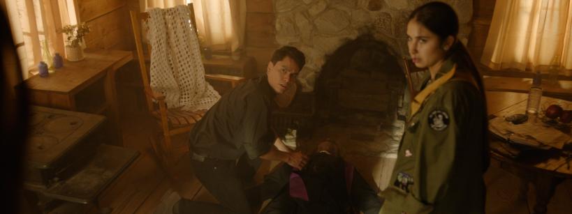 A young man kneels beside an unconscious or injured person lying on the floor of a rustic cabin, while a woman in a scout uniform stands nearby looking concerned. The room is warmly lit with wooden walls and furniture.