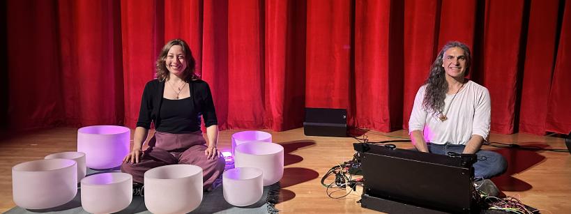 Two people sit on stage in front of a red curtain. One is surrounded by white crystal singing bowls, and the other sits beside electronic equipment. Both are smiling, seated on the floor under stage lighting.