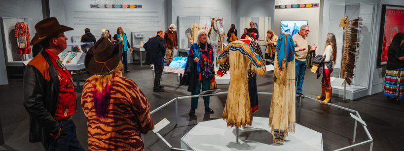 A group of people view traditional Indigenous clothing and artifacts displayed in a well-lit museum exhibit, with informational plaques and showcases on the walls and vibrant patterns featured on the garments.