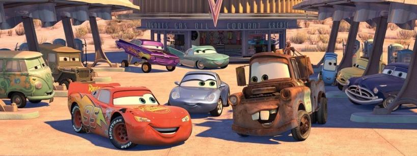 Animated cars with expressive faces gather at a gas station in a desert town. The group includes a red race car, a rusty tow truck, a blue car, a green van, and more, all smiling and interacting in front of a building.