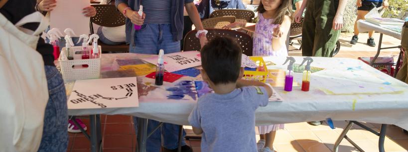 Children and adults gather around a table under a red canopy, engaging in arts and crafts activities with paints and paper at an outdoor event. People chat and work together in a lively, sunny environment.