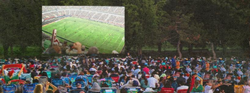 A large crowd sits outdoors on lawn chairs facing trees, watching a projected image of a stadium with a soccer game. The scene suggests a public outdoor viewing event.