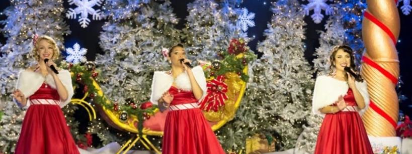 Three women in matching red dresses and white shawls sing on stage decorated with Christmas trees, gifts, and lights. The festive backdrop includes gold elements and snowflakes, evoking a cheerful holiday atmosphere.
