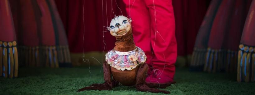 A marionette puppet dressed in a frilly outfit is controlled by strings, standing on green grass with a person in red pants behind it, in front of a stage with pink curtains.