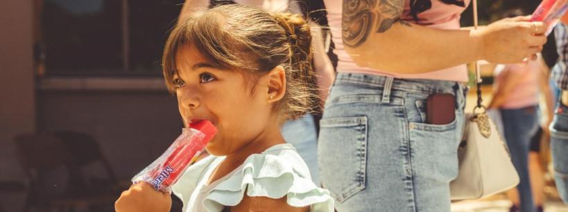 A young girl with brown hair in a bun enjoys a red popsicle outdoors. Adults stand nearby, one with a tattooed arm and another with keys clipped to their jeans. It appears to be a sunny day.