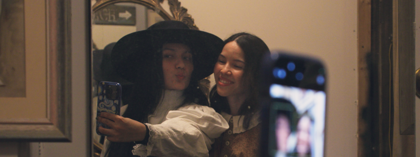 Two women pose for a selfie in front of a mirror; one wears a black hat and white blouse, and the other smiles in a tan vest. A phone held up in the foreground captures their reflection.