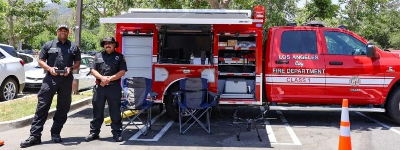 Two uniformed firefighters stand beside a red Los Angeles Fire Department command vehicle with open compartments displaying equipment. Folding chairs and traffic cones are set up nearby in a sunny outdoor area.
