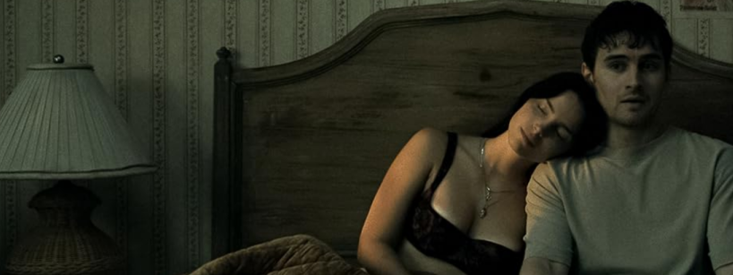 A woman sleeps with her head on a man's shoulder as they sit on a bed in a dimly lit room with patterned wallpaper and a lamp on the nightstand.