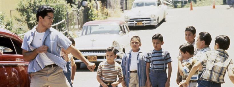 A teenage boy strikes a dramatic pose in front of a group of young boys on a sunlit street with vintage cars parked nearby. The boys watch him with curious and surprised expressions.