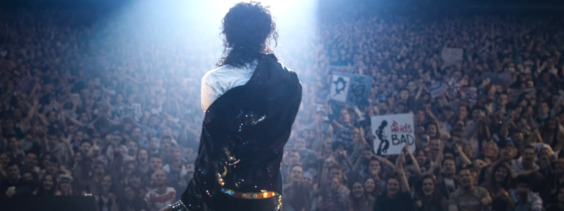 A performer stands on stage facing a large, cheering crowd with spotlights shining down. Some audience members hold up signs, including one that says "Bad.