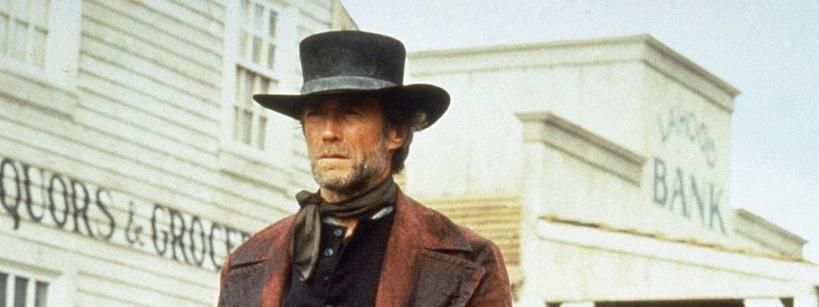 A man in a dark coat and tall black hat stands in front of old wooden buildings, including one marked “BANK,” with a western town setting and a serious expression on his face.
