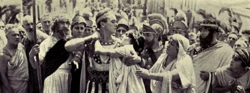 A group of people in ancient Roman attire surrounds a central figure being attacked, while others look on in shock or distress. The scene appears dramatic and intense, set on a crowded street.