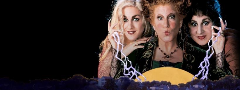 Three women in witch costumes make mischievous faces, holding glowing lightning above a town at night with a large yellow moon behind them.