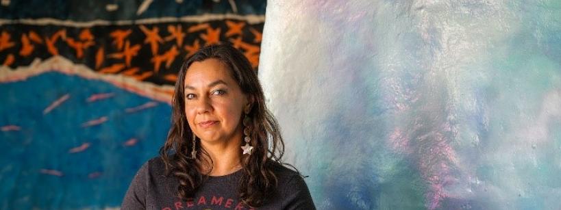 A woman with long brown hair and tattoos sits in front of colorful, abstract artwork. She wears a “Dreamers & Schemers” t-shirt and star earrings, smiling gently at the camera.