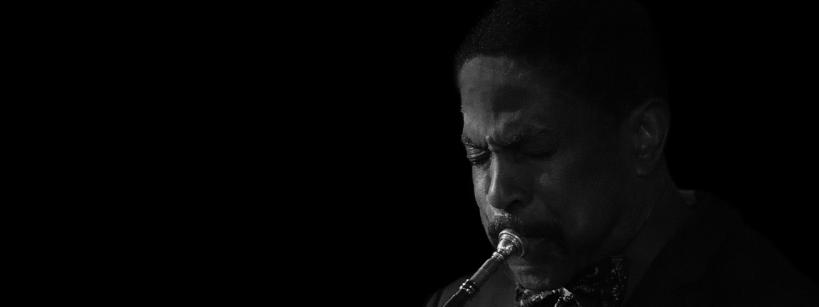 A man in a suit and bow tie plays a trumpet with concentration. The background is black, highlighting the musician and his instrument. The image is in black and white.