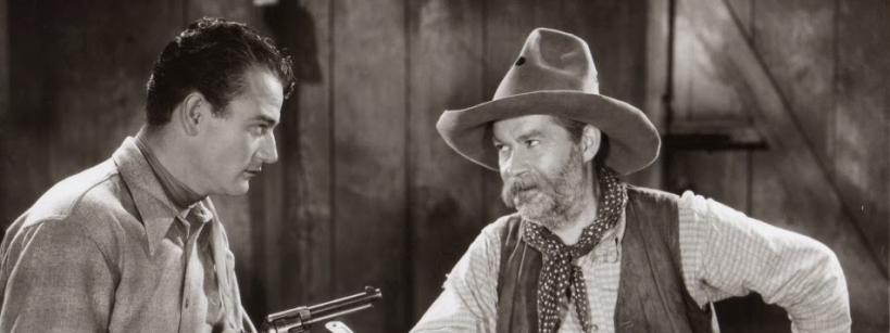 Two men in Western attire sit at a table with plates of food. One man points a revolver while the other, wearing a cowboy hat and scarf, looks at him. The setting appears to be an old wooden cabin.