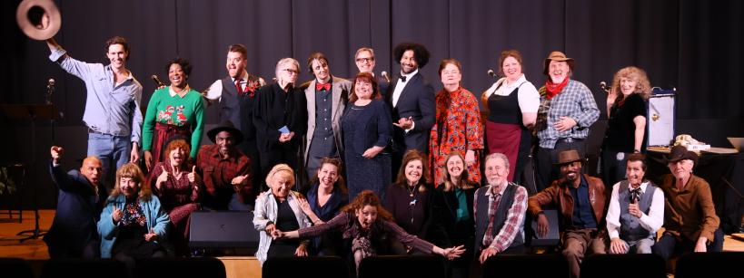 A group of about 25 adults pose and smile on a stage, some standing and some sitting, in front of a curtain. One person holds a hat in the air. The atmosphere is lively and cheerful.