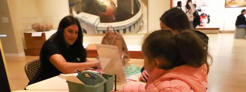 A young girl in a pink coat draws with colored pencils at a table, guided by an adult woman, in a brightly lit art gallery or museum space. Other children are also engaged in the activity.