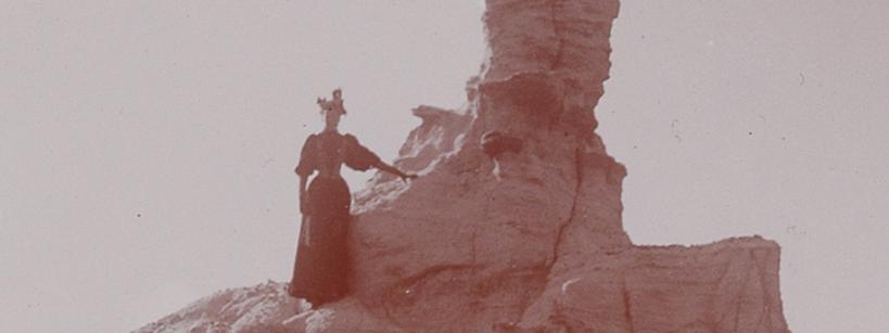 woman in 19th century clothing stands on steep hill next to rock formation