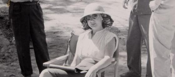 A woman wearing a light-colored hat and dress sits on a chair outdoors, holding an open book. Three men stand around her, their faces out of frame. The scene appears to be from an earlier era.